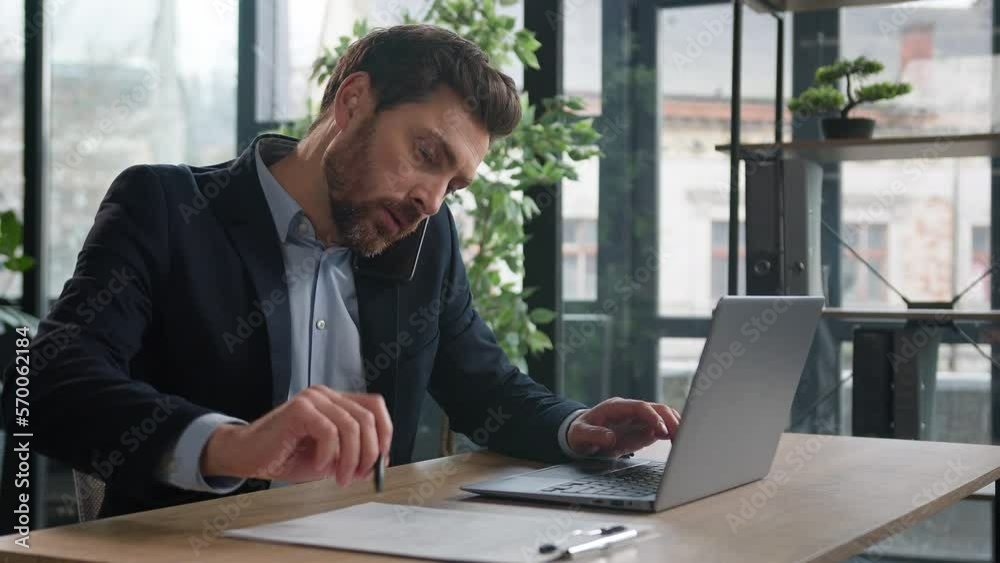 Busy successful caucasian adult businessman in office typing on laptop talking to client on mobile phone writing notes explain condition multitasking man manager communicate on cellphone in workplace