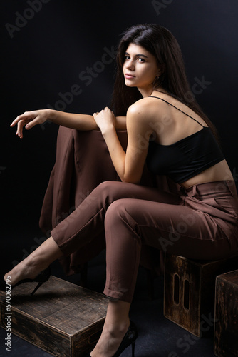 Classic studio portrait of a young brunette dressed in a black top and formal suit, who is sitting on a chair against a black background. © Budjak Studio