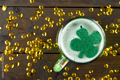 Image of beer glass, clover and gold confetti on wooden background