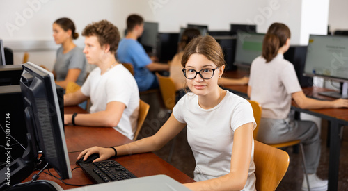 Portrait of positive smart girl in glasses sitting at desk in computer class, smiling and looking at camera.
