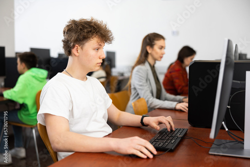 Teenage boy sitting at table and using computer during computer science lesson. His teacher and classmates standing and looking at monitor.
