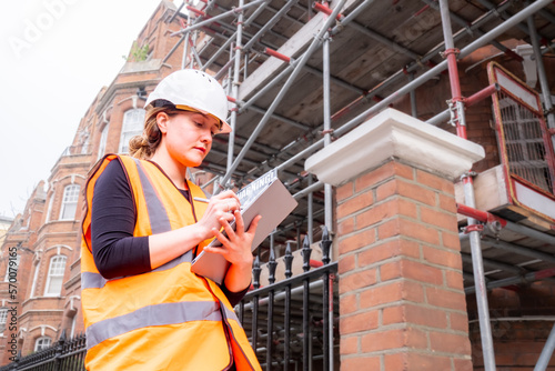 Chartered civil engineer woman writing on a tablet with electronic pen, hard hat and orange personal protective equipment, inspection, touchscreen, technology and innovation in construction site