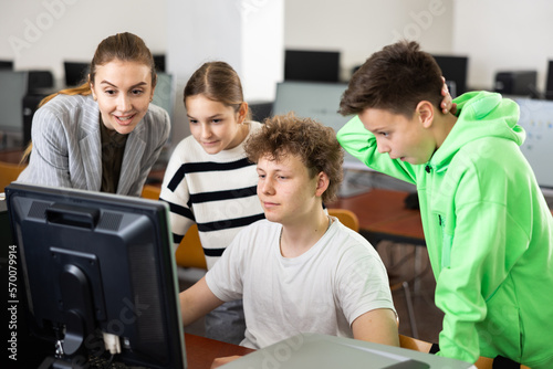 Teenage boy sitting at table and using computer during computer science lesson. His teacher and classmates standing and looking at monitor.