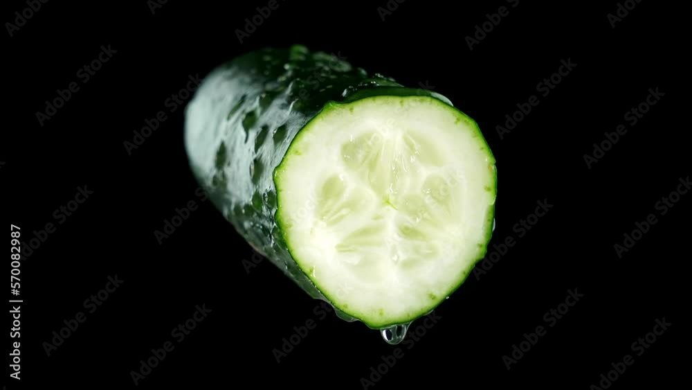 Half of tasty fresh green broken cucumber with clear water drops turns slowly on deep black background extreme close view