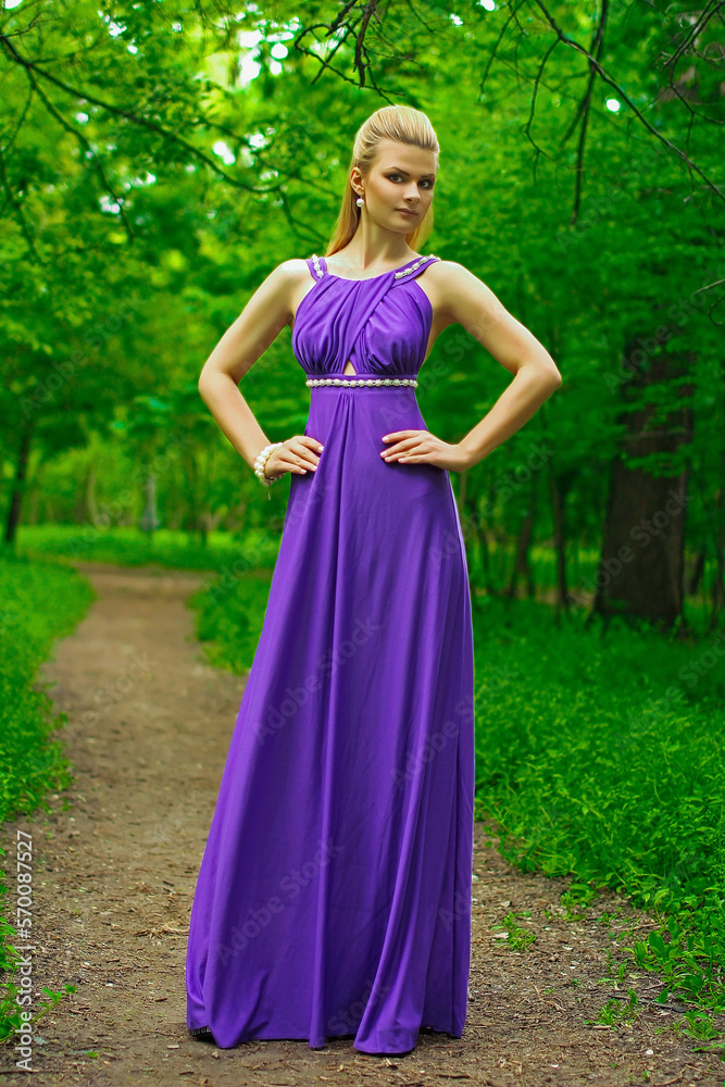 blonde beautiful natural girl with blonde hair in a lilac dress in nature, in the park, in the garden by a tree, with makeup and hairstyle without a bra