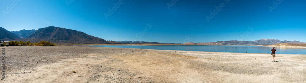 Panoramic view of Crowley Lake and the surrounding mountains, during a sunny day. A person walks near the edge of the lake. Panoramic view of Crowley Lake and the surrounding mountains, during a sunny day. A person walks near the edge of the lake.