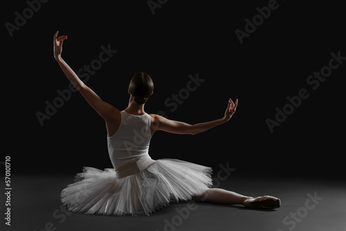Young ballerina sitting on split against black background, back view
