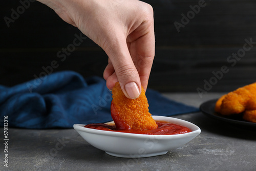 Woman dipping delicious chicken nugget into ketchup at grey table, closeup