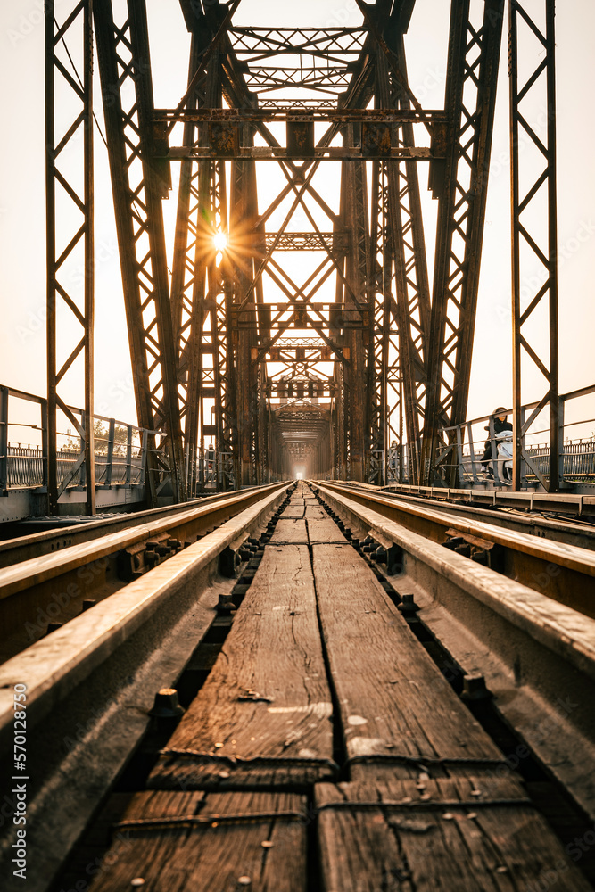 Train tracks lead downwards over a long metal bridge. There is a warm evening atmosphere.
