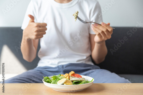 Asian teenage boy smile and happy with bowl of fresh salad  sit on sofa in a room. Healthy eating.