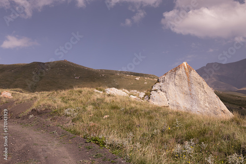 Mountain landscape - huge grey pyramid boulder with orange lichen on roadside of country road in sunlight in sunny summer day with blue sky, dry meadow and ridges away. Travel in Dagestan mountain.