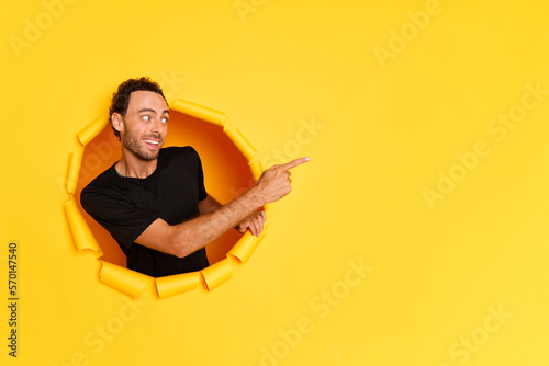 Portrait shot of impressed young man points index finger away to copy space looks aside with amazed expression shows promotional content dressed in black t-shirt poses through paper hole