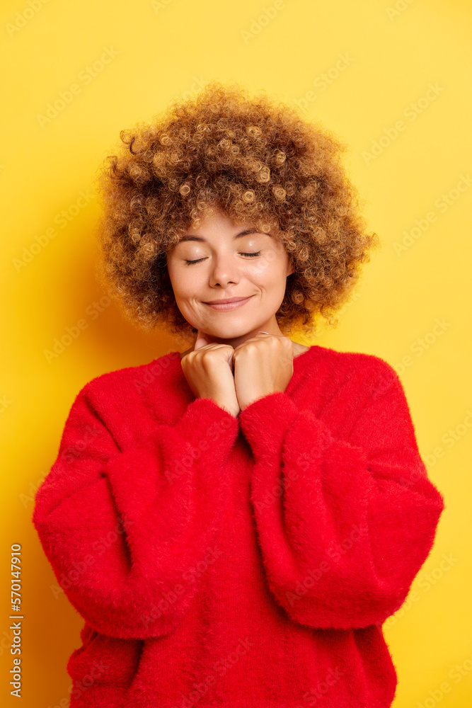 Portrait of kind lovely curly-haired European woman holding hands in prayer with closed eyes in a wish indoor studio shot isolated over yellow background Portrait of kind lovely curly-haired European woman holding hands in prayer with closed eyes in a wish indoor studio shot isolated over yellow background