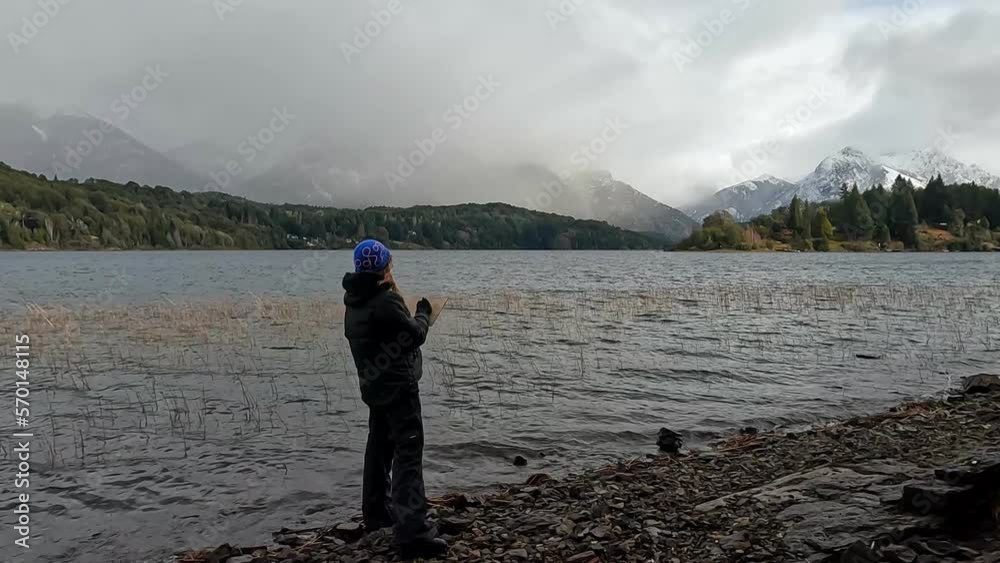 A female artist is sketching the landscape beside a lake in Patagonia. The scene is atmospheric. The mountains in the distance are snowcapped. A female artist is sketching the landscape beside a lake in Patagonia. The scene is atmospheric. The mountains in the distance are snowcapped.
