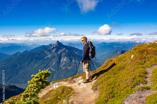 Adventurous athletic male hiker standing on a hiking trail on top of a rugged mountain in the Pacific Northwest with jagged mountains in the background. 