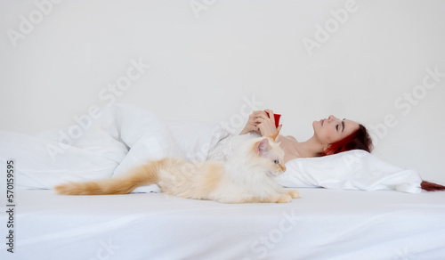 Portrait of an attractive, contented, young, sexy red-haired woman lying relaxed in white bed enjoys her coffee in a red cup and her cat lies beside her
