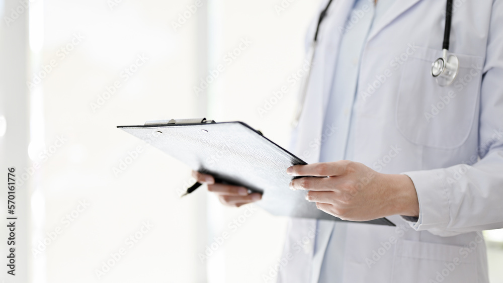 Female doctor with a stethoscope stands in the examination room, holding a medical clipboard. Female doctor with a stethoscope stands in the examination room, holding a medical clipboard.