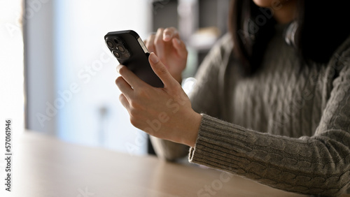 Close-up image of an Asian woman in cozy sweater sitting at her desk and using her smartphone.