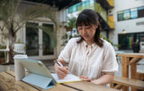 Young asian girl with freckles smiling and studying online with a tablet, notes and pen outdoors