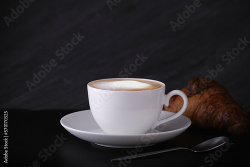 White cup with cappuccino on a dark background. Italian breakfast on a black background. Cappuccino with croissant on a black background.
