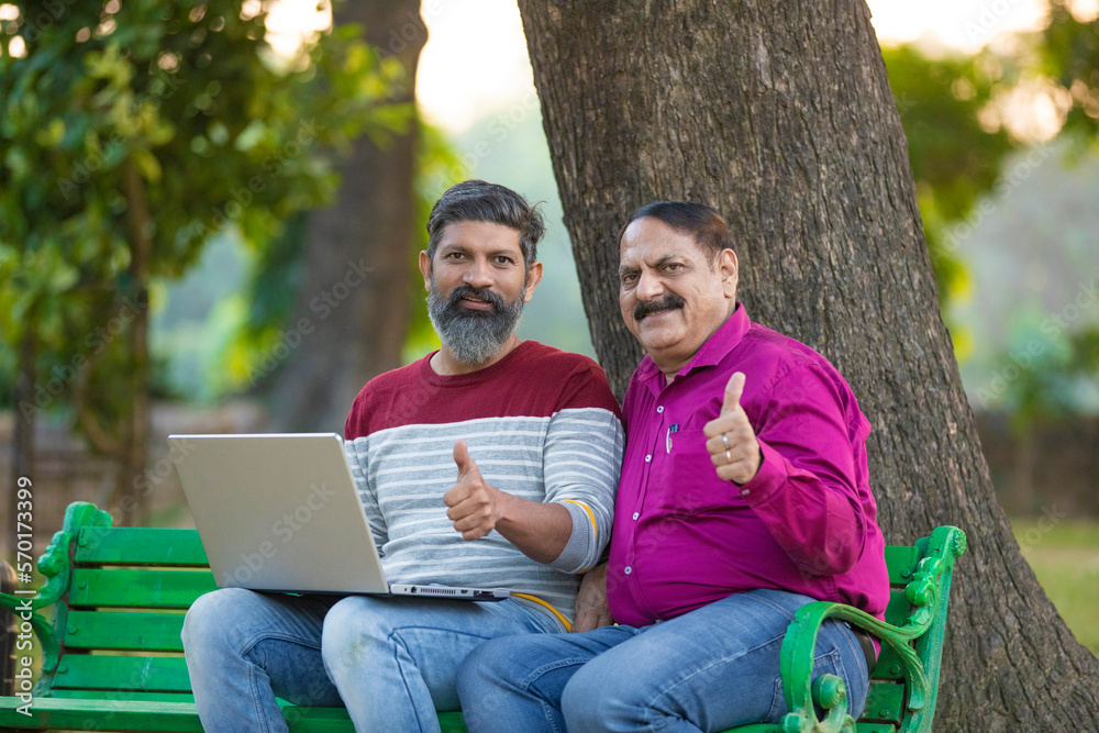 Two indian people using laptop and showing thumps up at park.