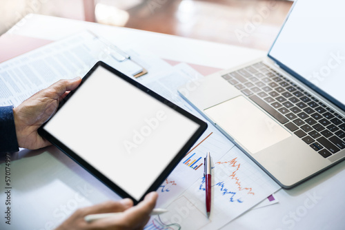 Asian businessman holding tablet computer use laptop with blank screen at workplace and use data charts in documents to analysis investment plan in future