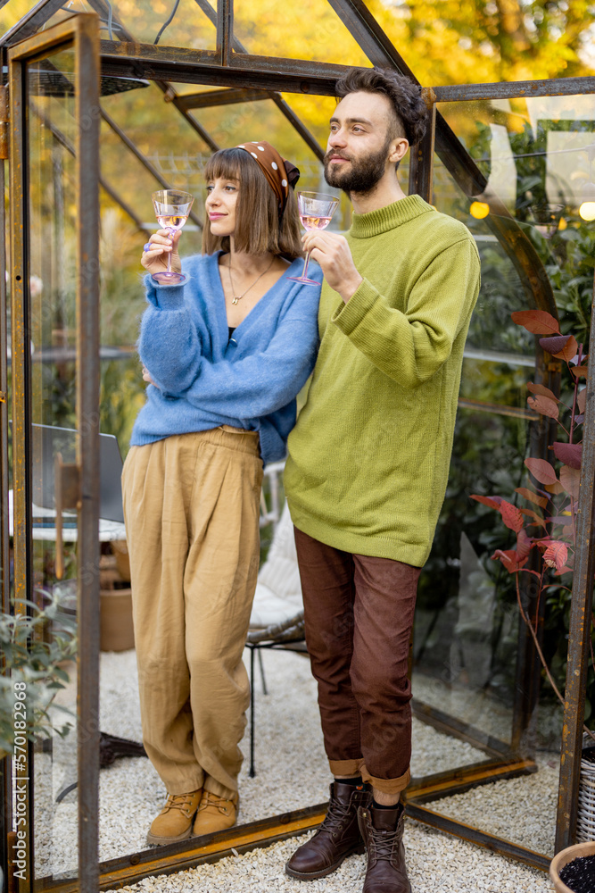 Fototapeta premium Young stylish couple talk and drink wine, spending leisure time together near glass house in garden. Man and woman have romantic eve time with alcohol drink at backyard