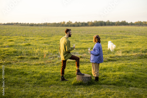Young stylish couple hang out together  talking and drinking wine on green field during sunset. Spending autumn time outdoors