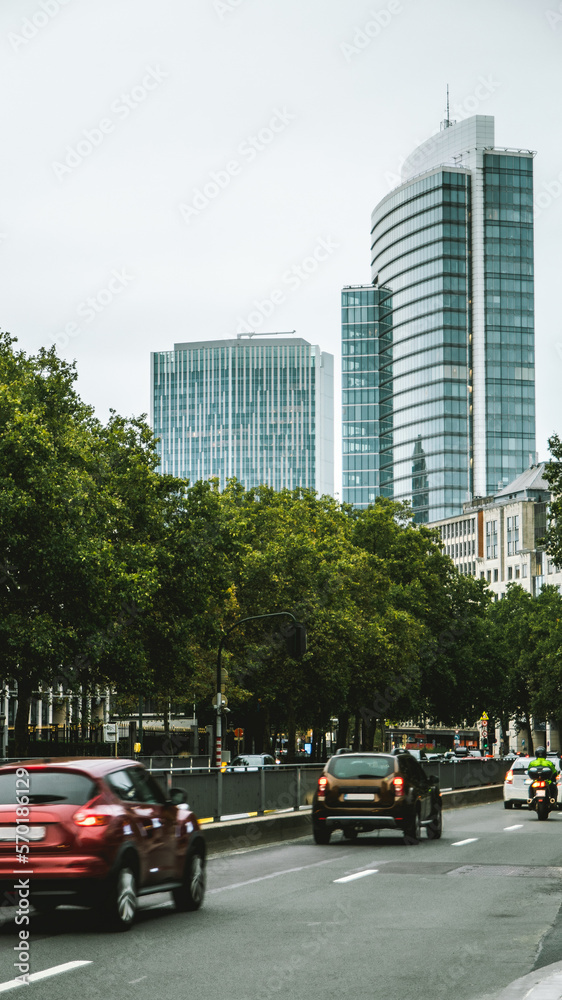 Obraz premium traffic on a road with trees on a sunny day and blue glass skyscrapers of downtown at the background