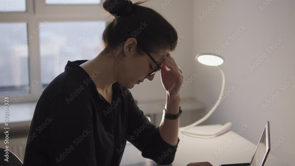 Exhausted young woman with hair bun and in eyeglasses feeling headache, rubbing forehead during