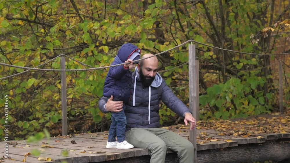 Bearded father and son are sitting on river bridge and looking at water flow. They talk, spend leisure time together. A man and 2 years old boy sit on a wooden bridge. Day vacation on river outdoors.