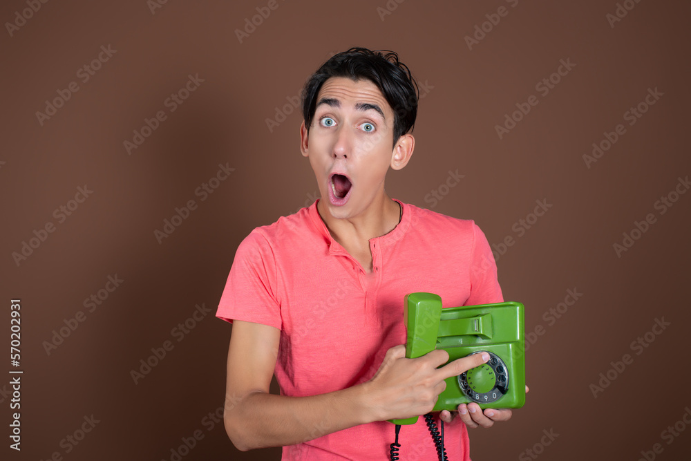 Funny young guy is talking on a landline phone. Student in a pink t-shirt on a brown background. A man with a retro telephone. Funny young guy is talking on a landline phone. Student in a pink t-shirt on a brown background. A man with a retro telephone.
