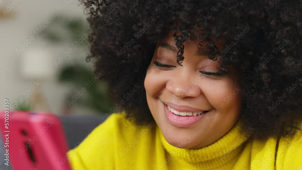 Close up view of smiling young black latin woman with curly hair having fun using mobile phone at home. Cheerful afro american female using app on cellphone relaxing on sofa. Technology concept. Close up view of smiling young black latin woman with curly hair having fun using mobile phone at home. Cheerful afro american female using app on cellphone relaxing on sofa. Technology concept.
