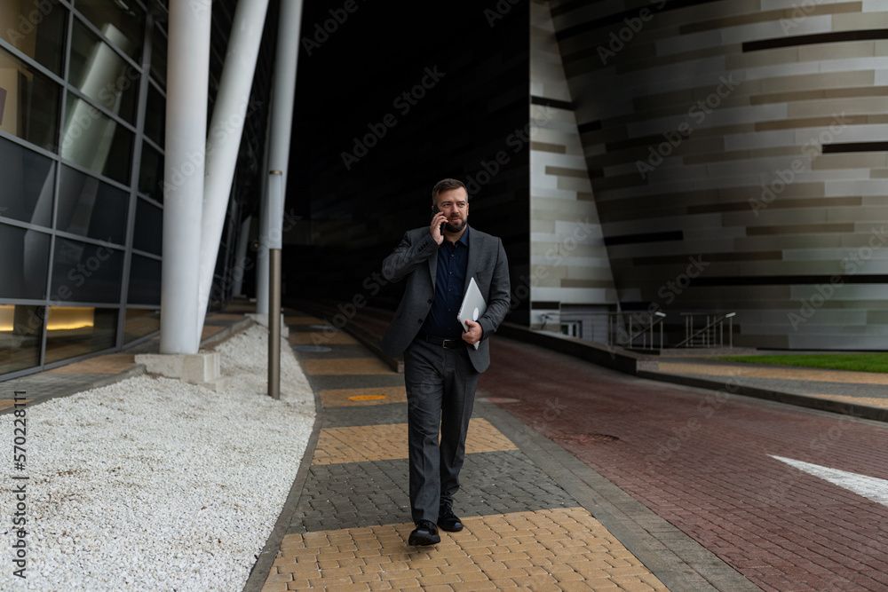 mature business man in a business suit talking on the phone with a laptop in his hands walking along the building mature business man in a business suit talking on the phone with a laptop in his hands walking along the building