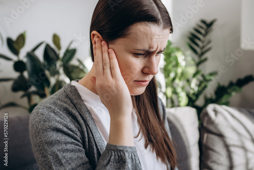 Close up portrait of upset young girl holding painful ear, suddenly feeling strong ache. Unhealthy caucasian woman 20s suffering from painful otitis sitting on sofa at home. Health problems concept