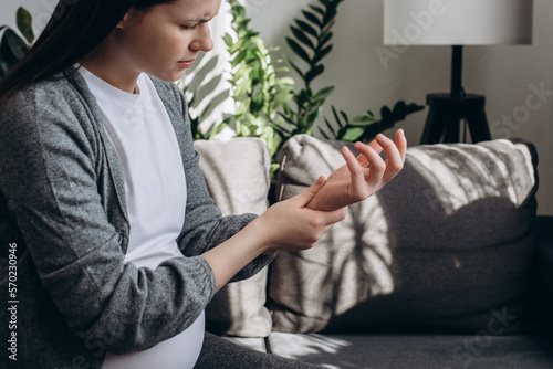 Selective focus of sad pregnant woman suffering pain on hands and fingers, arthritis inflammation. Upset caucasian female has pain in wrist sitting on couch at home. Health care and medical concept photo