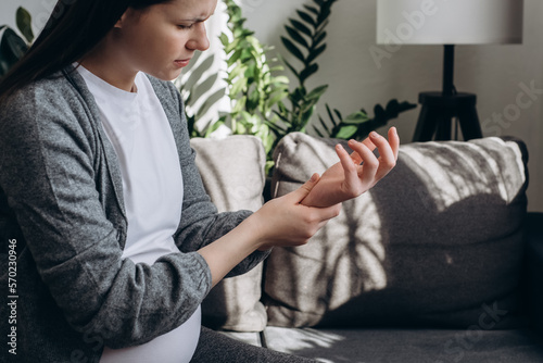 Selective focus of sad pregnant woman suffering pain on hands and fingers, arthritis inflammation. Upset caucasian female has pain in wrist sitting on couch at home. Health care and medical concept