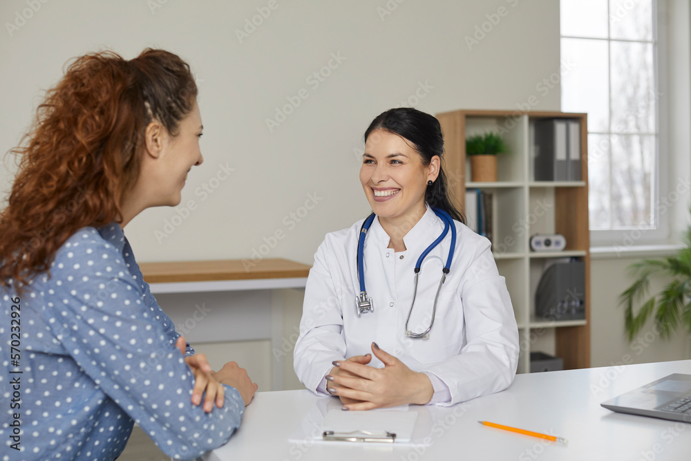 Beautiful smiling caring female doctor having pleasant conversation with patient in medical office. Woman in white coat and with stethoscope sitting at table smiling at young female patient. Beautiful smiling caring female doctor having pleasant conversation with patient in medical office. Woman in white coat and with stethoscope sitting at table smiling at young female patient.
