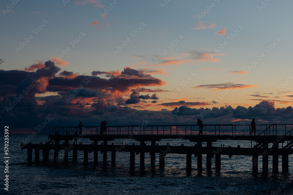 Sunset on the coast, people are enjoying and taking photos of it on the pier. Clouds in the background painted in orange and little magenta colors