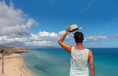 Hombre con camiseta verde quitándose el sombrero de paja frente a la playa de arena blanca y el mar turquesa de Jandia en el centro turístico de Fuerteventura en las Islas Canarias.