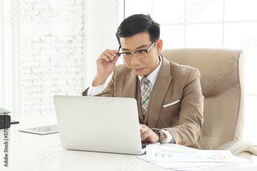 Attractive cheerful asian classy CEO businessman  in suit and with eyeglasses using laptop while looking at the camera, Asian boss middle age. CEO business man in formal clothing wearing spectacles
