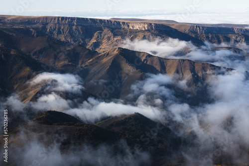 Photo of a scenic view of hills in the lowland and clouds crawling between hills under sunlight and clouds shadows on the plateau Bermamyt. There are trees in autumn colors on the hills.