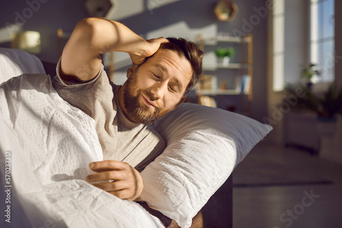 Attractive bearded young man just woke up and did not have time to open his eyes. Smiling cute guy, lying in his bed at home in the bedroom, holding his head with his hand. Good morning, beginning day