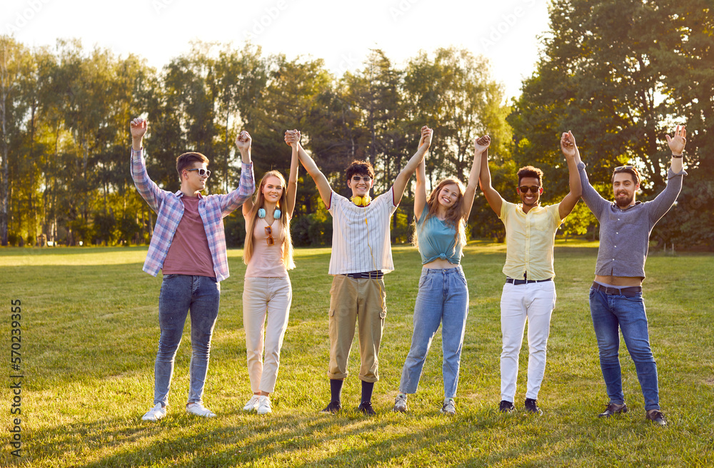 Team unity. Portrait of cheerful friendly multiracial friends holding hands standing in row in summer park. Men and women stand with arms raised and smiling at camera. Cohesion concept. Team unity. Portrait of cheerful friendly multiracial friends holding hands standing in row in summer park. Men and women stand with arms raised and smiling at camera. Cohesion concept.