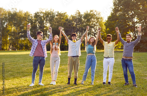 Team unity. Portrait of cheerful friendly multiracial friends holding hands standing in row in summer park. Men and women stand with arms raised and smiling at camera. Cohesion concept.