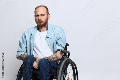 A man in a wheelchair looks at the camera, with tattoos on his arms sits on a gray studio background, the concept of health is a person with disabilities