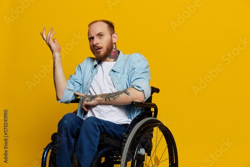 A man in a wheelchair problems with the musculoskeletal system looks at the camera is not satisfied, with tattoos on his arms sits on a yellow studio background, health concept man with disabilities