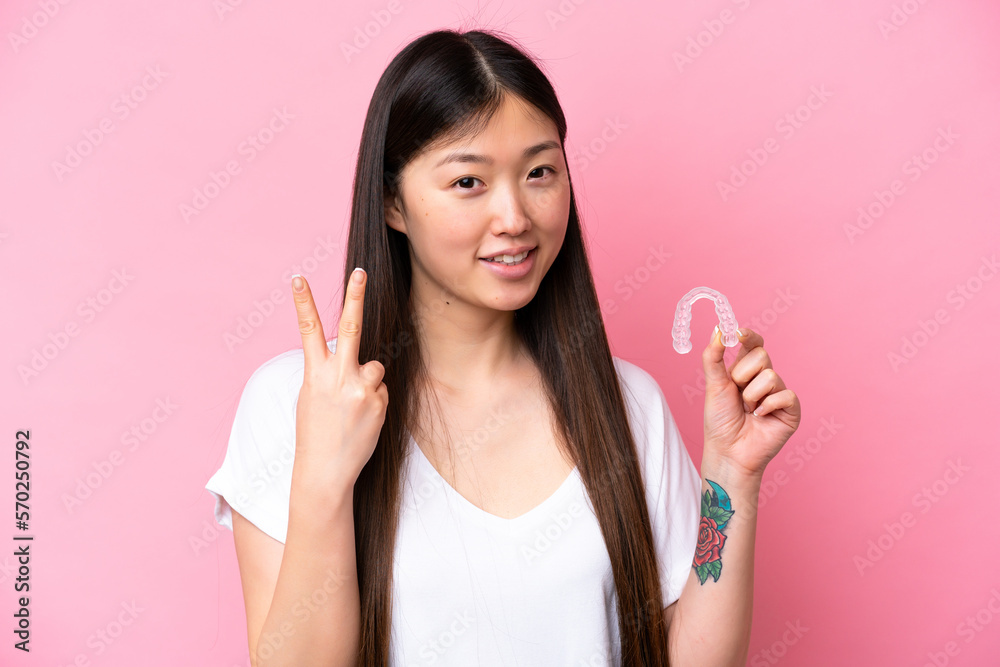 Young Chinese woman holding invisible braces isolated on pink background smiling and showing victory sign Young Chinese woman holding invisible braces isolated on pink background smiling and showing victory sign