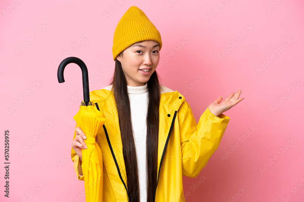 Young Chinese woman with rainproof coat and umbrella isolated on pink background extending hands to the side for inviting to come