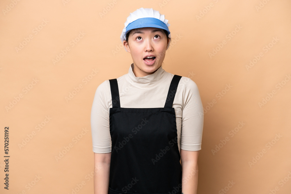 Young Chinese Fishmonger woman wearing an apron and holding a raw fish isolated on pink background looking up and with surprised expression